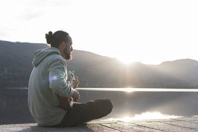 Man playing ukulele while sitting on promenade near lake