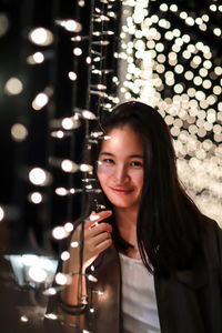 Portrait of smiling young woman by illuminated string lights at night