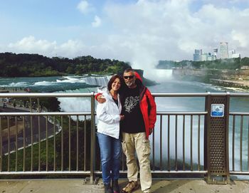 Portrait of couple standing on bridge over river against cloudy sky