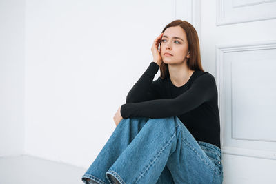 Young unhappy woman girl with long hair in jeans on floor in closed position by door at home