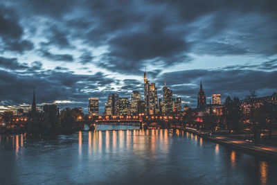 View of illuminated buildings by river against cloudy sky