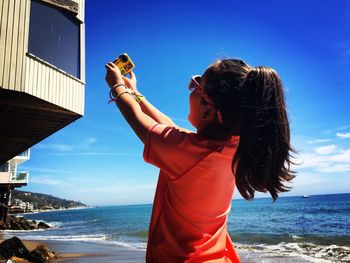 Low angle view of girl taking selfie from camera at beach against sky on sunny day