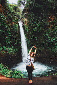 Happy woman standing with arms raised against waterfall in rainforest