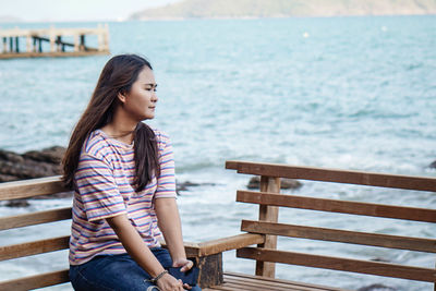 Young woman looking away while sitting on railing against sea