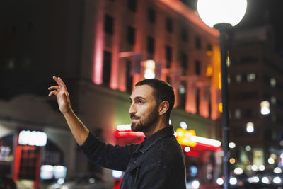 Side view of young man hailing taxi on city street at night