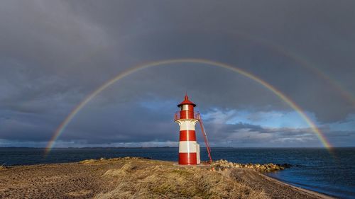 Lighthouse by sea against rainbow in sky