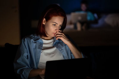 Serious focused woman freelancer working on laptop in dark at home looking at screen.