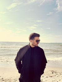 Portrait of young man standing at beach against sky during sunset