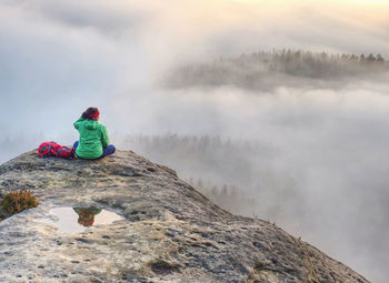 Green jacket woman pensively sitting on the edge of a rock and looking at the misty clouds.