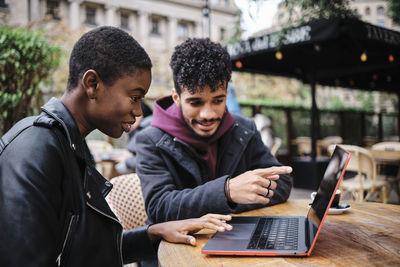 Man pointing toward laptop while sitting by friend at sidewalk cafe