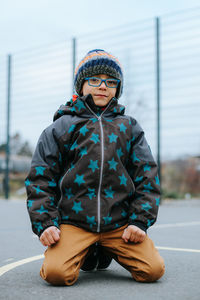 Portrait of cute boy wearing knit hat kneeling on road during winter