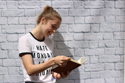 Young woman reading book while standing by wall