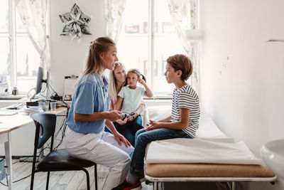 Female pediatrician with otoscope examining boy while family sitting in background at clinic