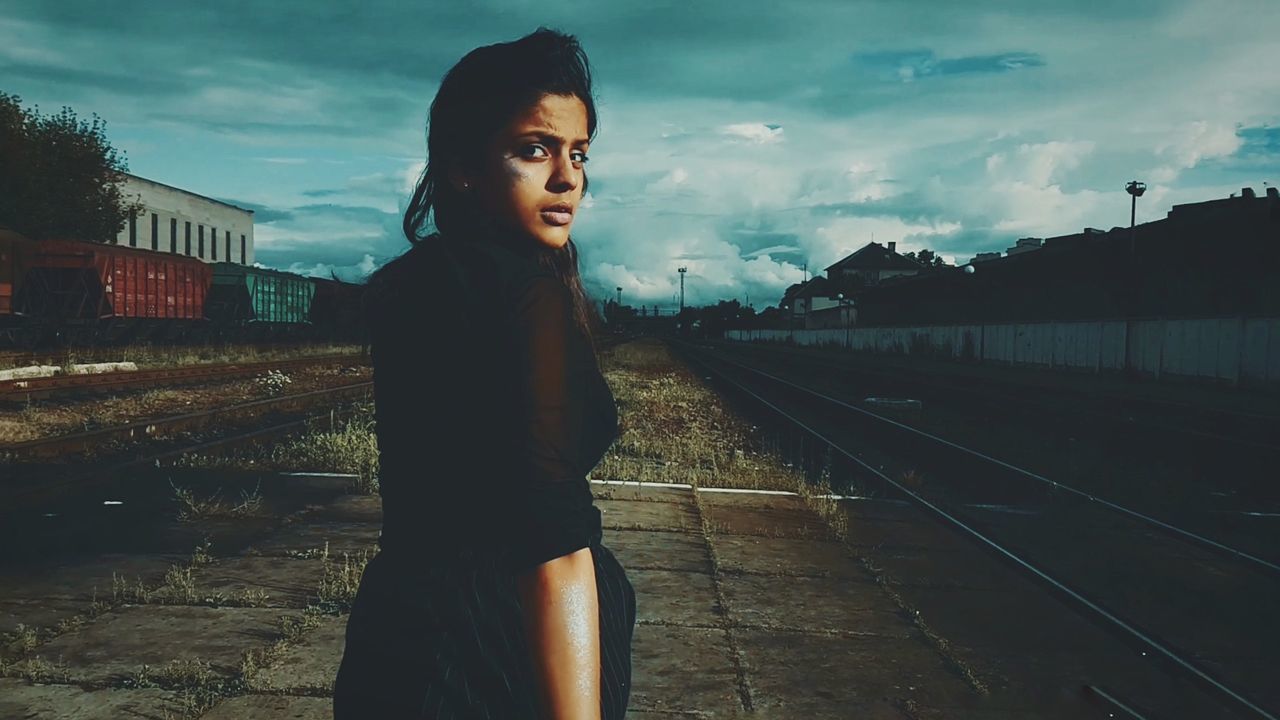 one person, sky, real people, young adult, rail transportation, standing, transportation, lifestyles, track, young women, leisure activity, railroad track, three quarter length, cloud - sky, casual clothing, nature, looking away, built structure, outdoors, waiting, contemplation, hairstyle