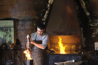 Young male blacksmith burning metal in fire at workshop