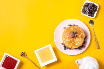 A stack of pancakes on pink plate with berries next to honey and strawberry jam on yellow background