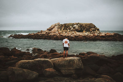 Rear view of man standing on rock by sea against sky