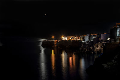 Illuminated buildings by sea against sky at night