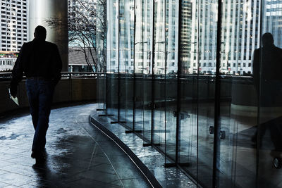 Rear view of man reflecting on glass wall while walking in passage