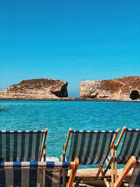 Deck chairs on rocks by sea against clear blue sky