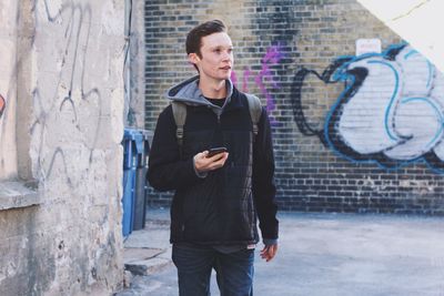 Young man holding mobile phone while looking away against wall