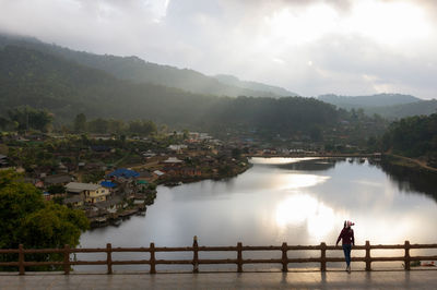 High angle view of woman standing on footbridge against lake and mountains during sunset