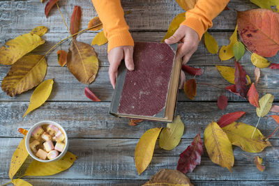 Cropped hand of woman holding book by autumn leaves on wooden table