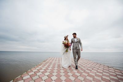 Newlywed couple walking on pier over lake against cloudy sky