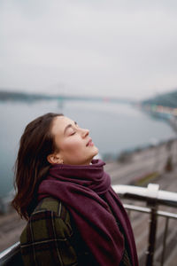 Beautiful calm young woman staying near river with closed eyes and expressing delight.