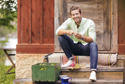 Smiling man cutting vegetables while sitting on steps outside cottage