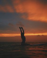 Silhouette young woman surfing on sea against cloudy sky during sunset