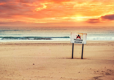 Rusty metallic warning sign on shore at beach against cloudy sky during sunset