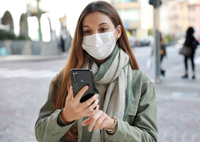 Smiling young woman wearing mask using smart phone in city