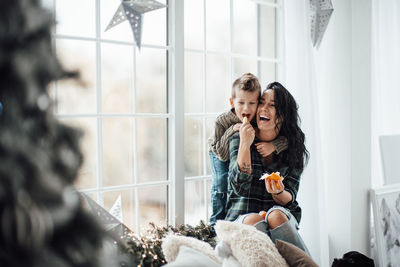 Laughing mother feeding orange to son sitting by window at home
