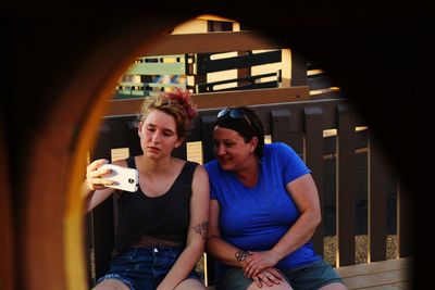 Female friends looking at mobile phone while sitting on bench seen through fence