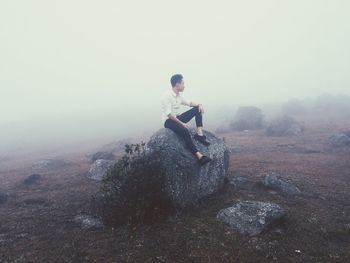 Man sitting on rock during foggy weather against sky