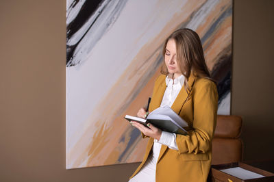 Young woman using digital tablet while standing against wall