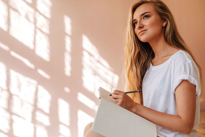 Beautiful woman writing in book while sitting against wall