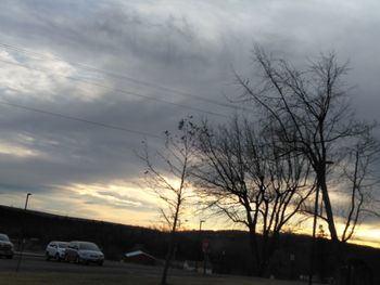 Silhouette bare trees by road against sky at sunset