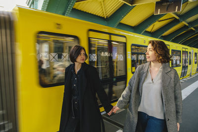 Lesbian couple hand hands walking by yellow train at railroad station