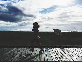 Side view of girl with kite walking on footpath against cloudy sky