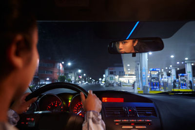 Back view of female driver reflecting in rear view mirror and driving modern automobile in city at night