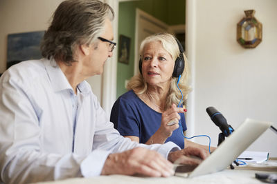 Senior woman wearing headphones and using microphone while discussing with male friend during vlogging at home