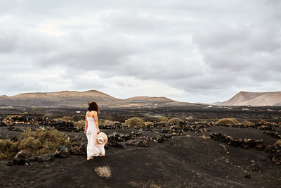 Unrecognizable woman in white dress carrying hat and walking on dry soil near bushes on cloudy day in waterless valley in fuerteventura, spain