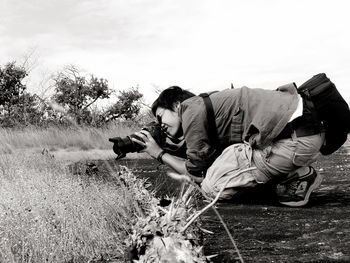 Side view of man photographing while crouching on field against sky
