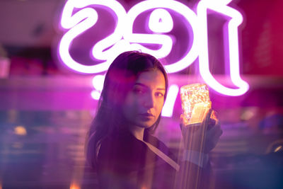 Portrait of young woman holding illuminated lighting equipment while standing against built structure at night