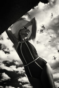 A portrait of a woman in a swimsuit, hat and sunglasses in summer on the riverbank against the sky