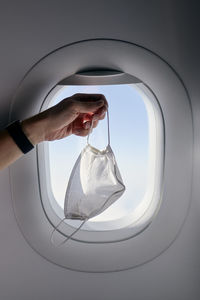 Hand holding protective face mask against airplane window. 