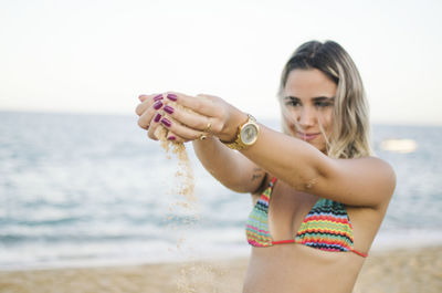 Smiling woman holding sand while standing at beach against sky