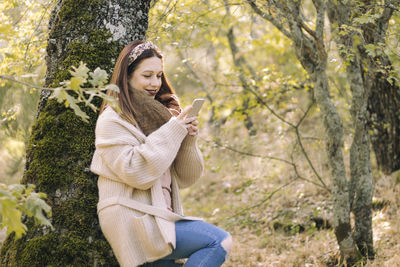 Smiling young woman, leaning against a tree, using her mobile phone